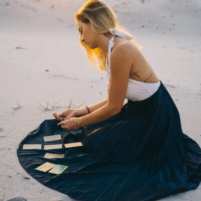 A woman sits on sand reading tarot cards during a desert sunset.